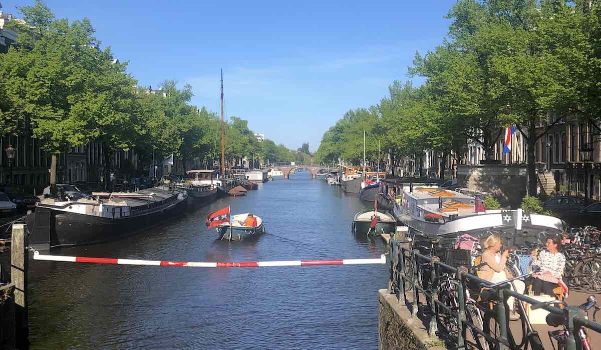 a boat going through the Keizersgracht in Amsterdam while two women enjoying the sunshine on the terrace with a drink in their hands
