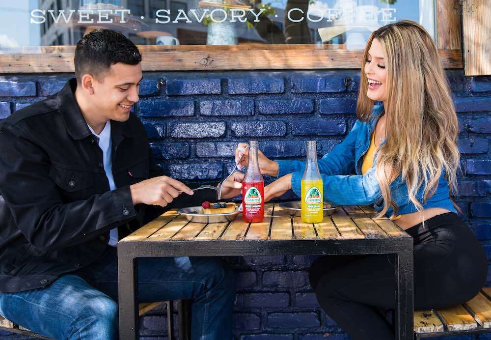 een man in een zwart jasje die uitgaat met een blonde vrouw in een blauw jasje die buiten op het terras zit te ontbijten.