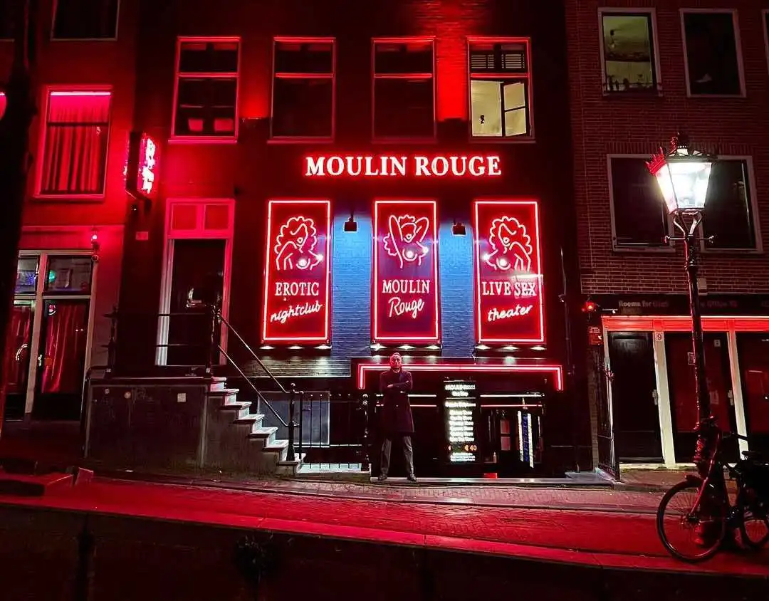The front of Moulin Rouge Amsterdam in the Red Light District with a bouncer in front of it surrounded by red light neon.