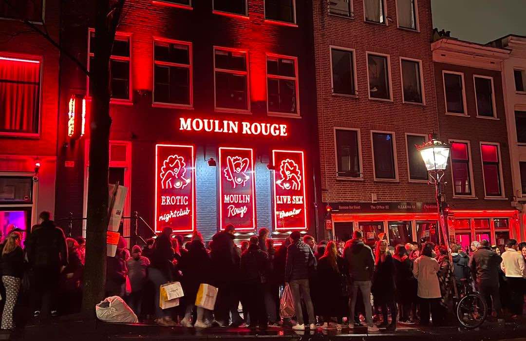 dozens of tourists walking by Moulin Rouge in Amsterdam Red Light District at night. Red-lit windows in the background