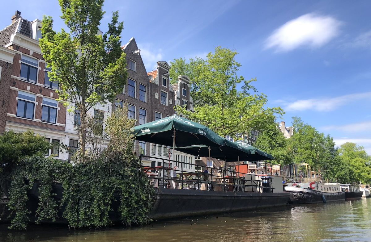 A leafy Jordaan canal in Amsterdam with a waterside cafe terrace under green umbrellas, historic gabled houses and moored houseboats